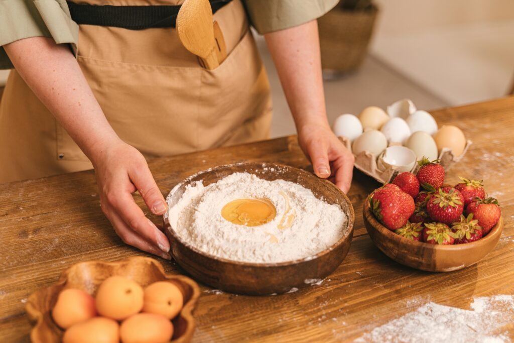 Hands preparing flour, eggs, and fruits for baking in the kitchen.