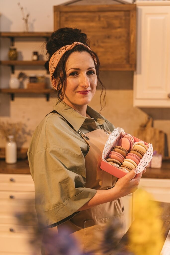 A woman stands in a rustic kitchen holding a box of colorful macarons with a warm smile.